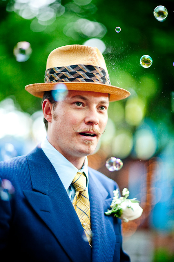 fun photo of the handsome groom with bubbles floating around him - groom is wearing a blue suit and a yellow tie with blue stripes and a straw derby style hat -photo by New Mexico based wedding photographers Twin Lens